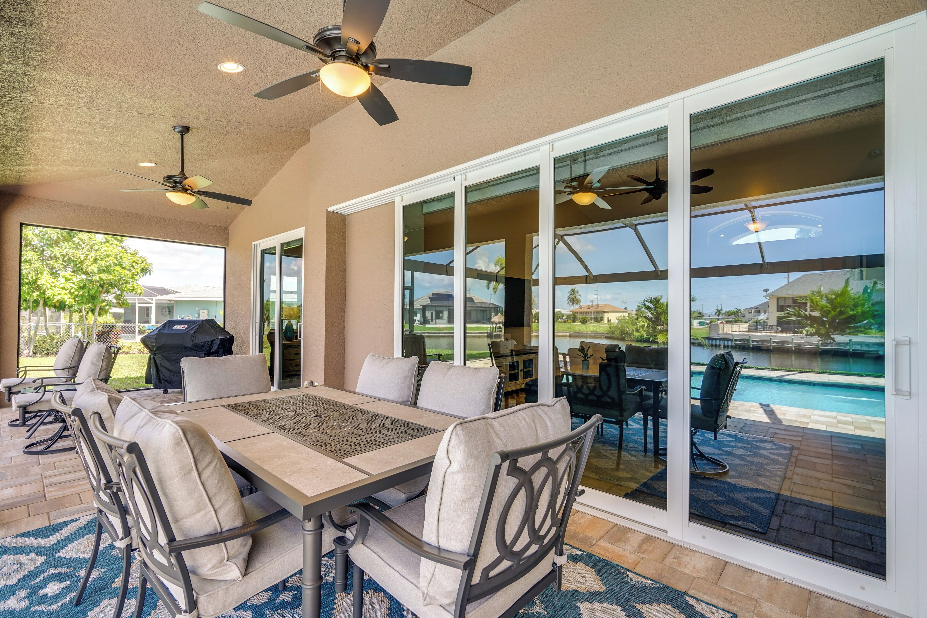 Un espace patio aménagé avec une table et des chaises, un ventilateur de plafond et une vue sur une piscine, visible à travers des portes vitrées.