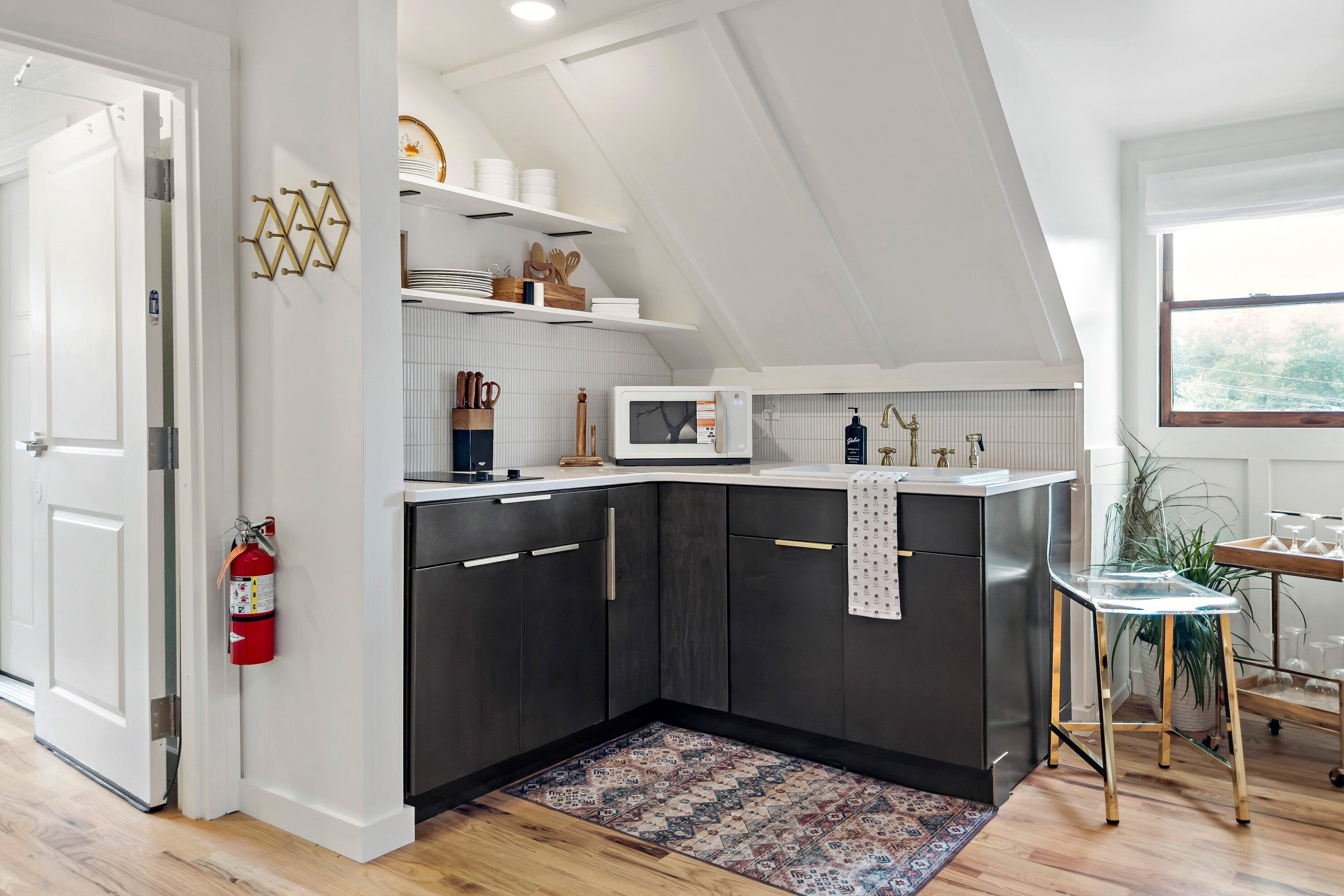 Une cuisine moderne avec des meubles de couleur sombre, un plan de travail blanc et un four à micro-ondes. On peut y voir un extincteur accroché au mur, une petite table avec des verres et une plante.