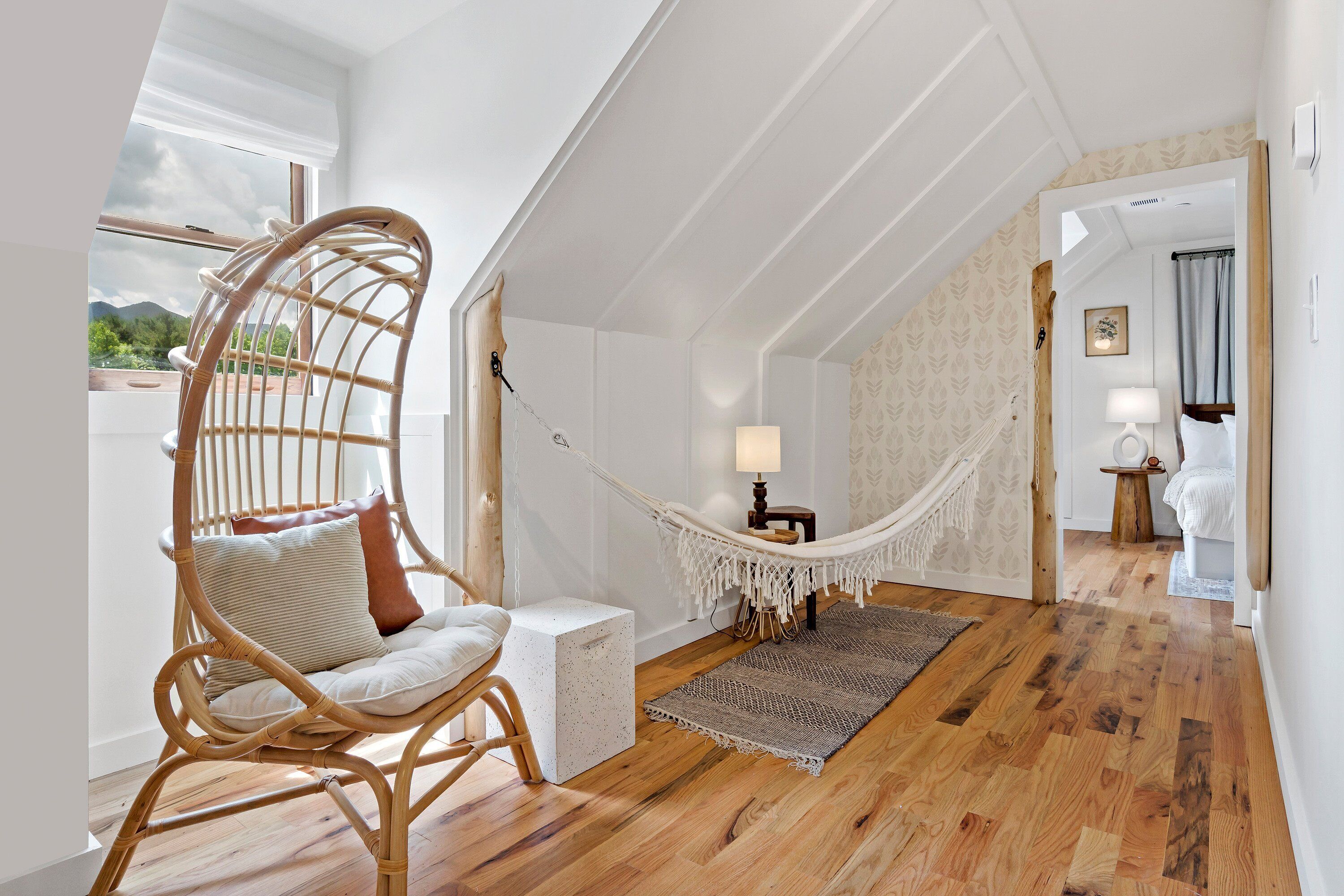 Una habitación con piso de madera, una silla de mimbre, una hamaca, una mesita con una lámpara y vistas al exterior.