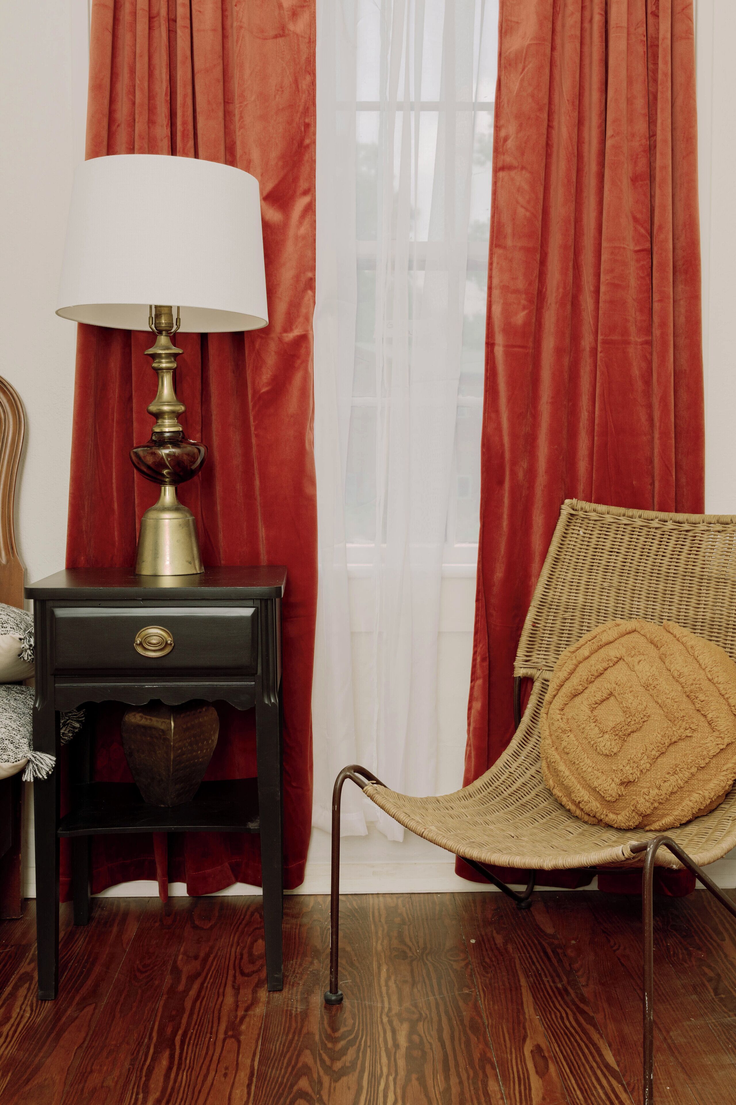 Une pièce avec une table de chevet en bois foncé, une lampe en laiton, un fauteuil en osier avec un coussin marron et des rideaux rouges.