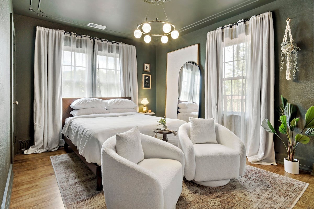 A bedroom with a large bed, two armchairs, a bedside table with a lamp, and a potted plant.