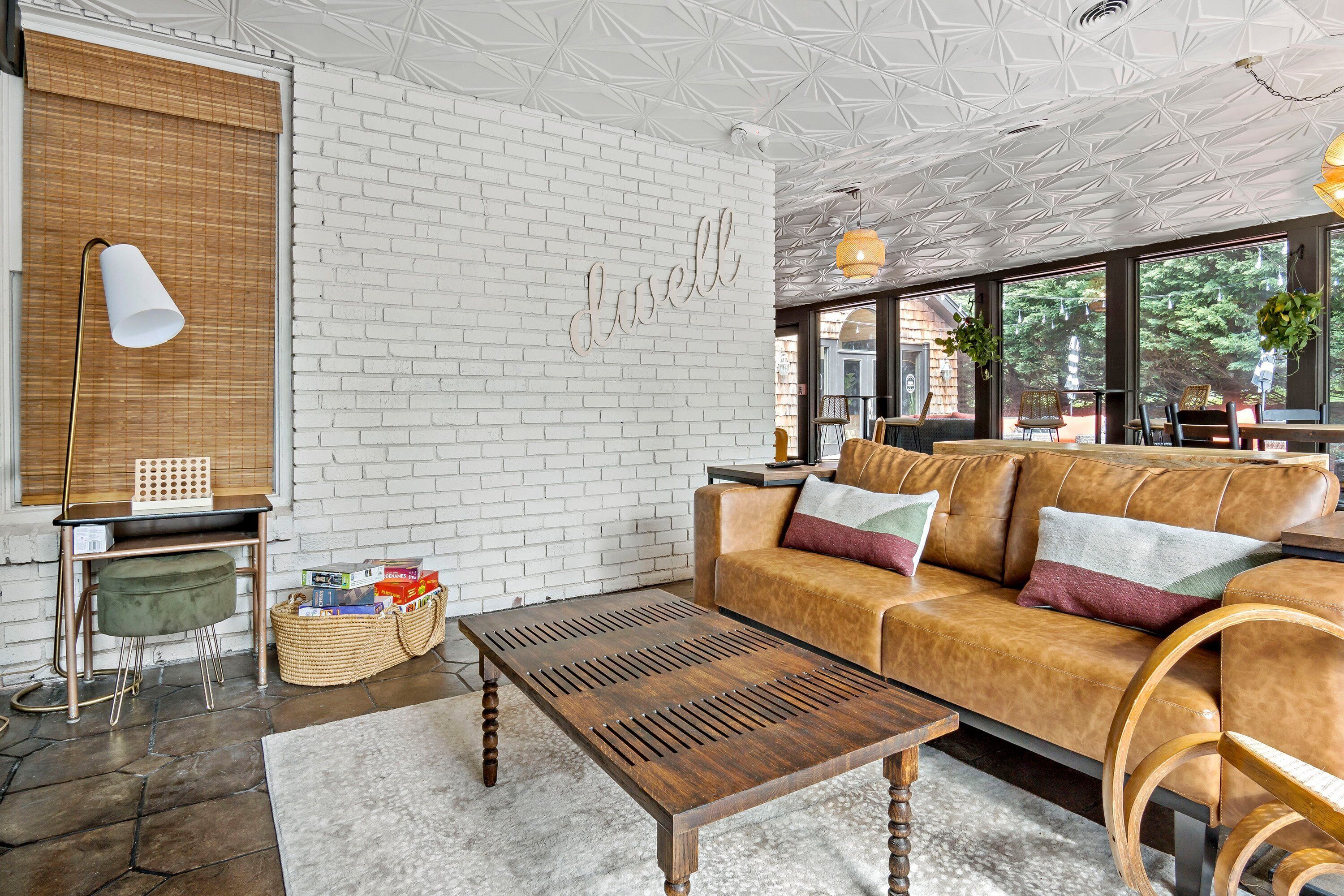 A modern living room with a white brick wall, a leather sofa, a wooden coffee table, and a large window with a view of greenery.