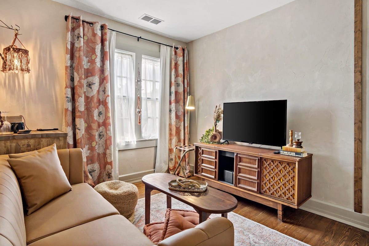 A living room with a sofa, a coffee table, a TV on a wooden cabinet, and a large window with curtains.