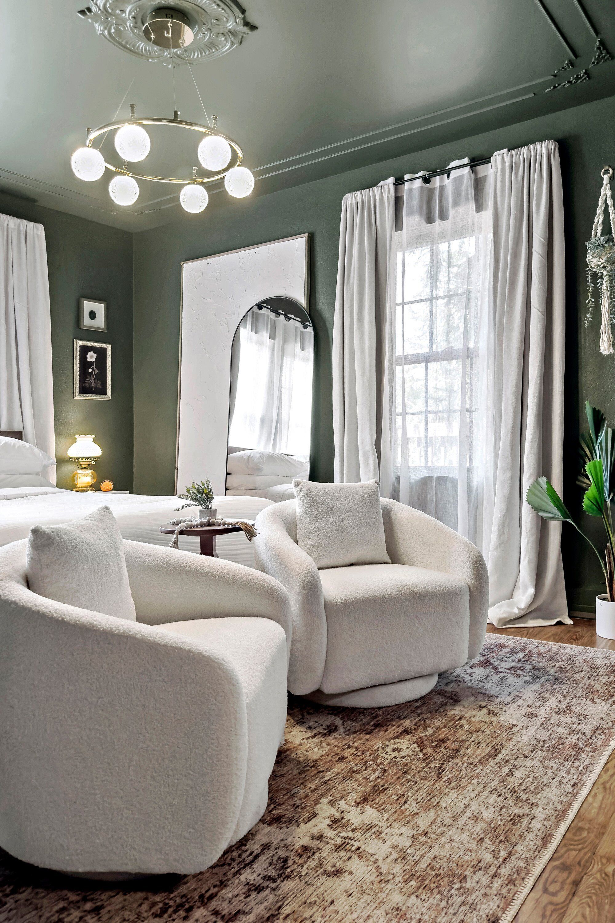 A room with a large mirror, two white armchairs, a small table with a lamp, and a large window with curtains.