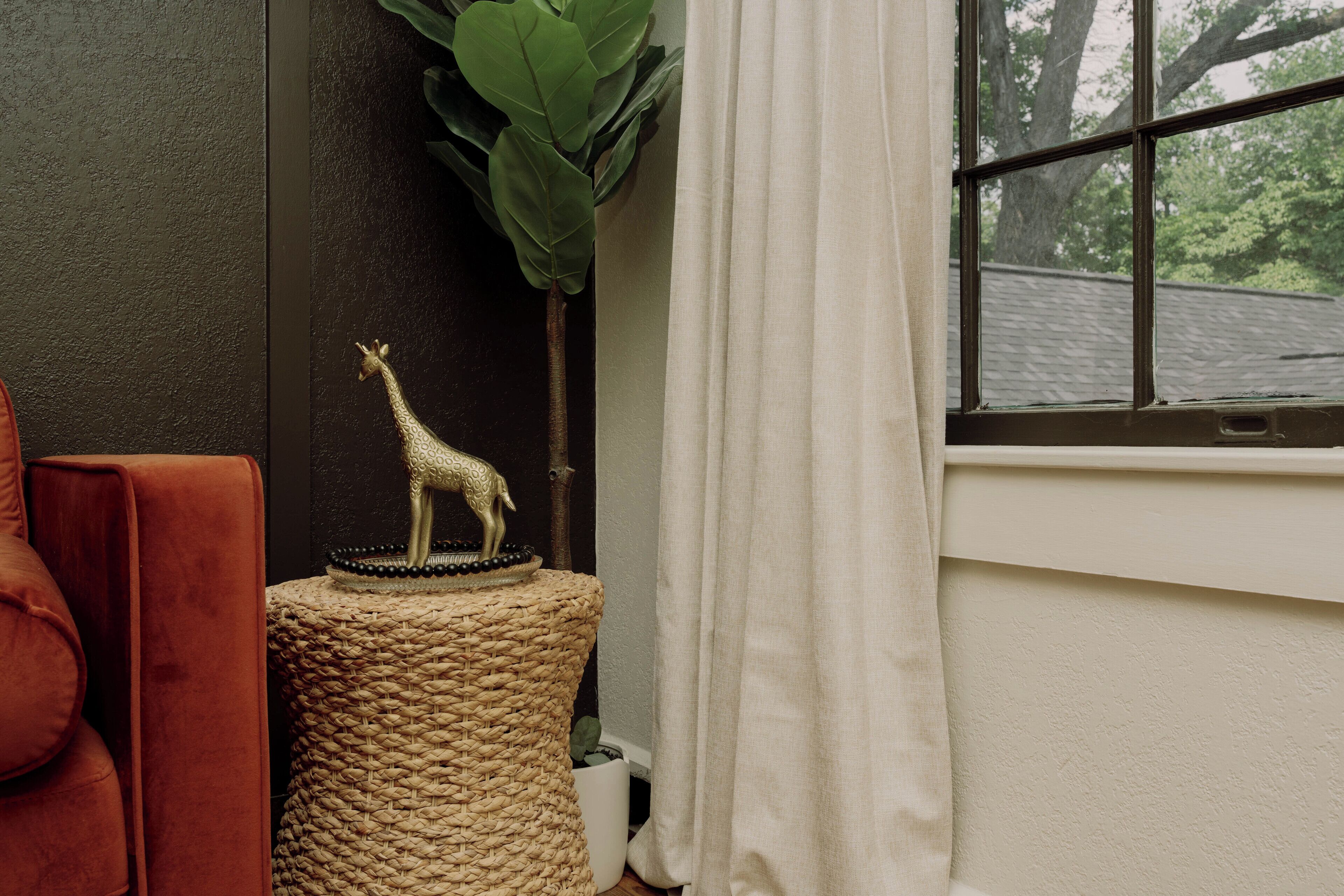 A room with a woven stool, a giraffe figurine, a red chair, and a window with a view of trees.