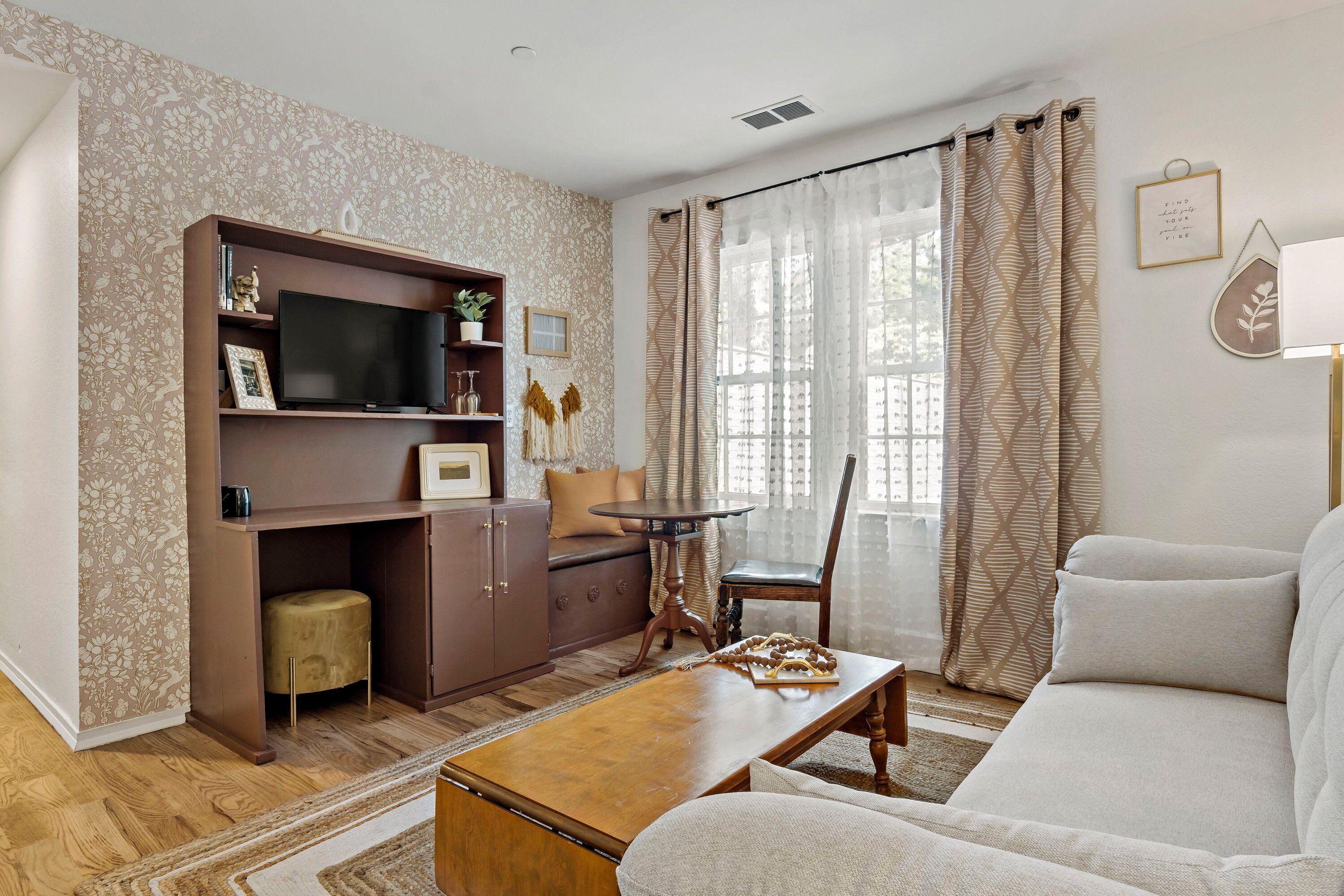 A living room with a sofa, a coffee table, a TV on a wooden stand, a small table with a chair, and patterned curtains.
