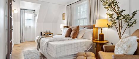 Un dormitorio con una cama grande, una mesita de noche con lámpara, una silla y una planta decorativa.