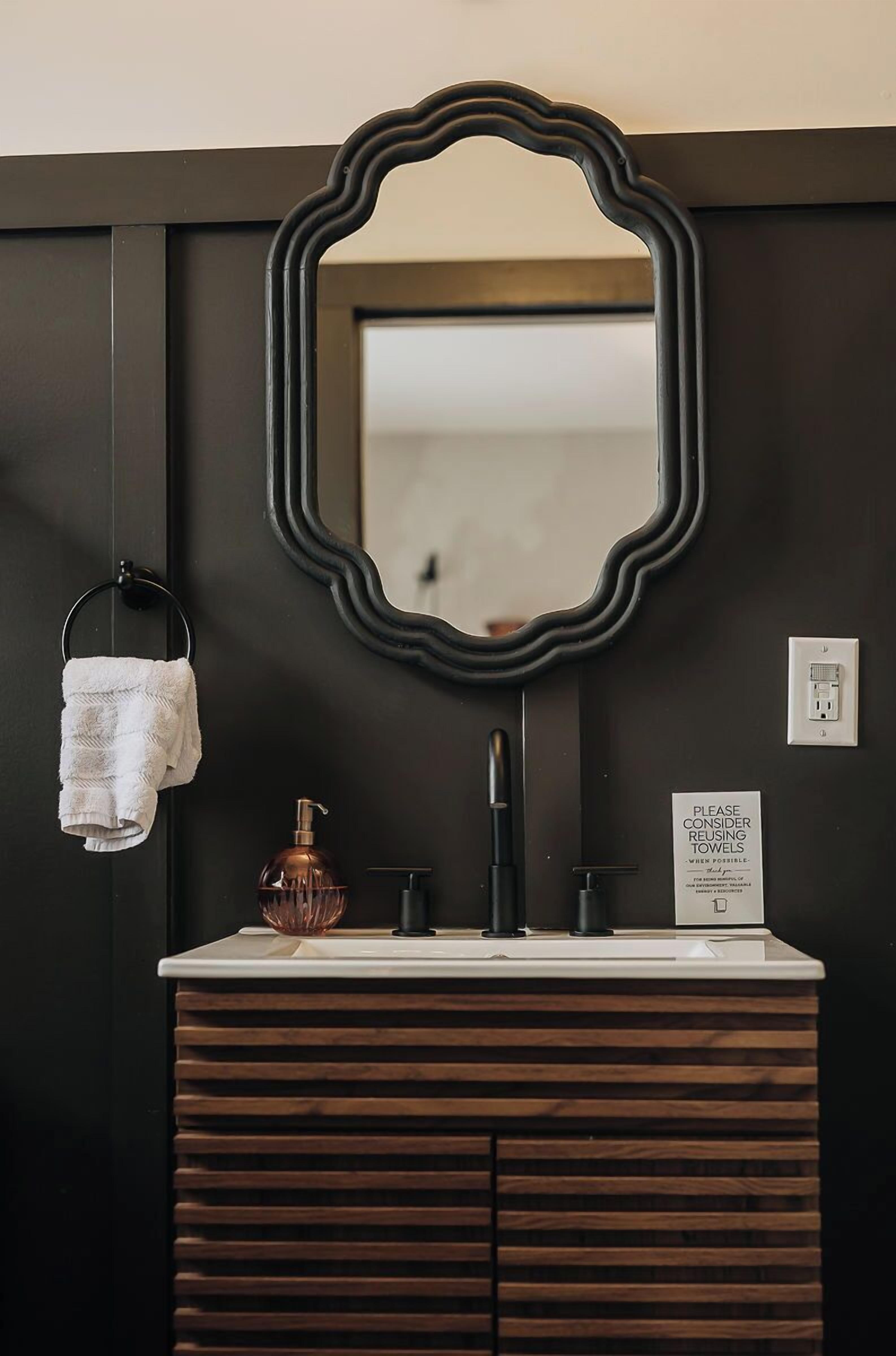 A bathroom with a dark wall, a wooden vanity, a mirror, a towel ring, a soap dispenser, and a sign about towel reuse.