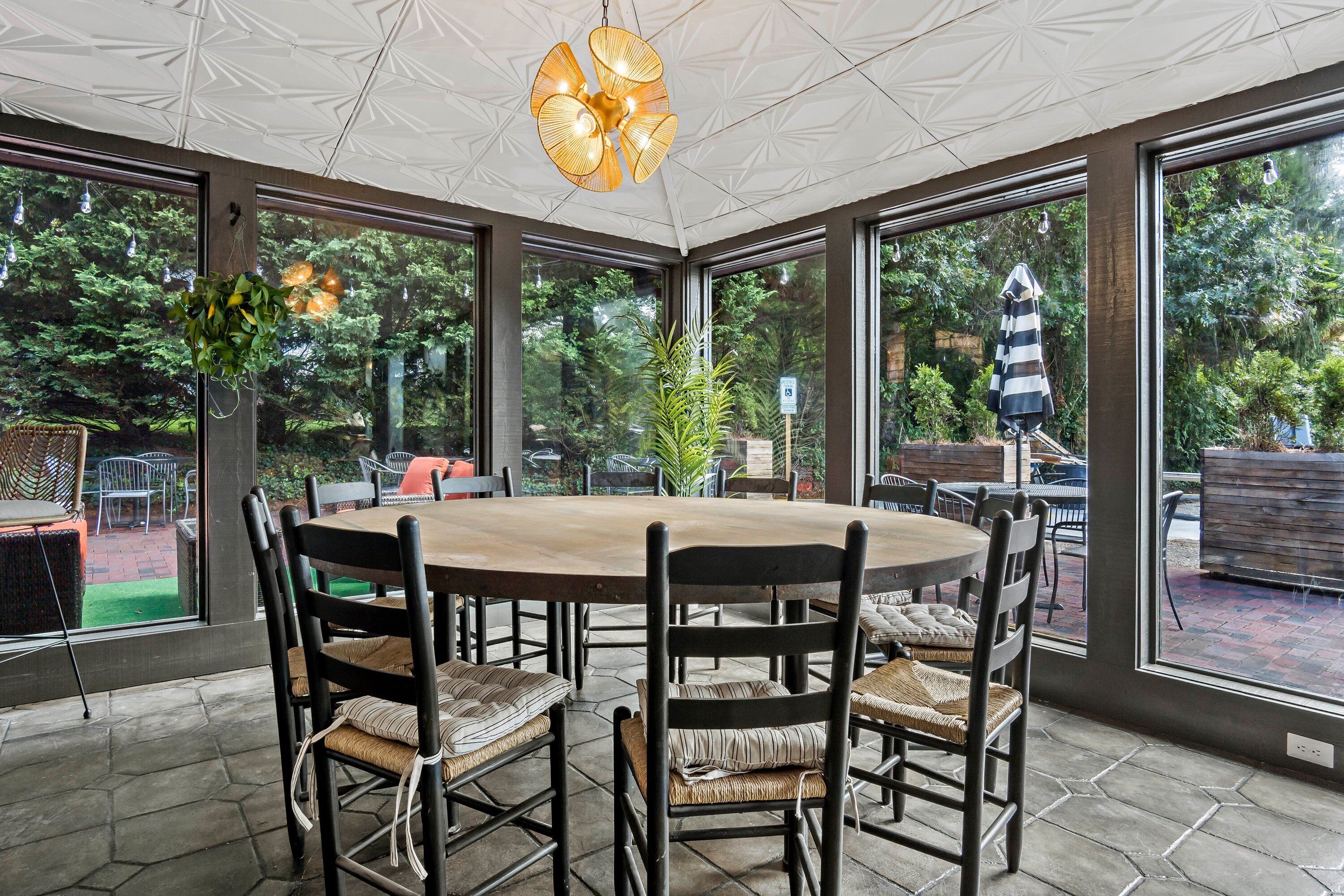 Ein überdachter Patio-Bereich mit einem runden Tisch und Stühlen, großen Fenstern und hängenden Pflanzen.