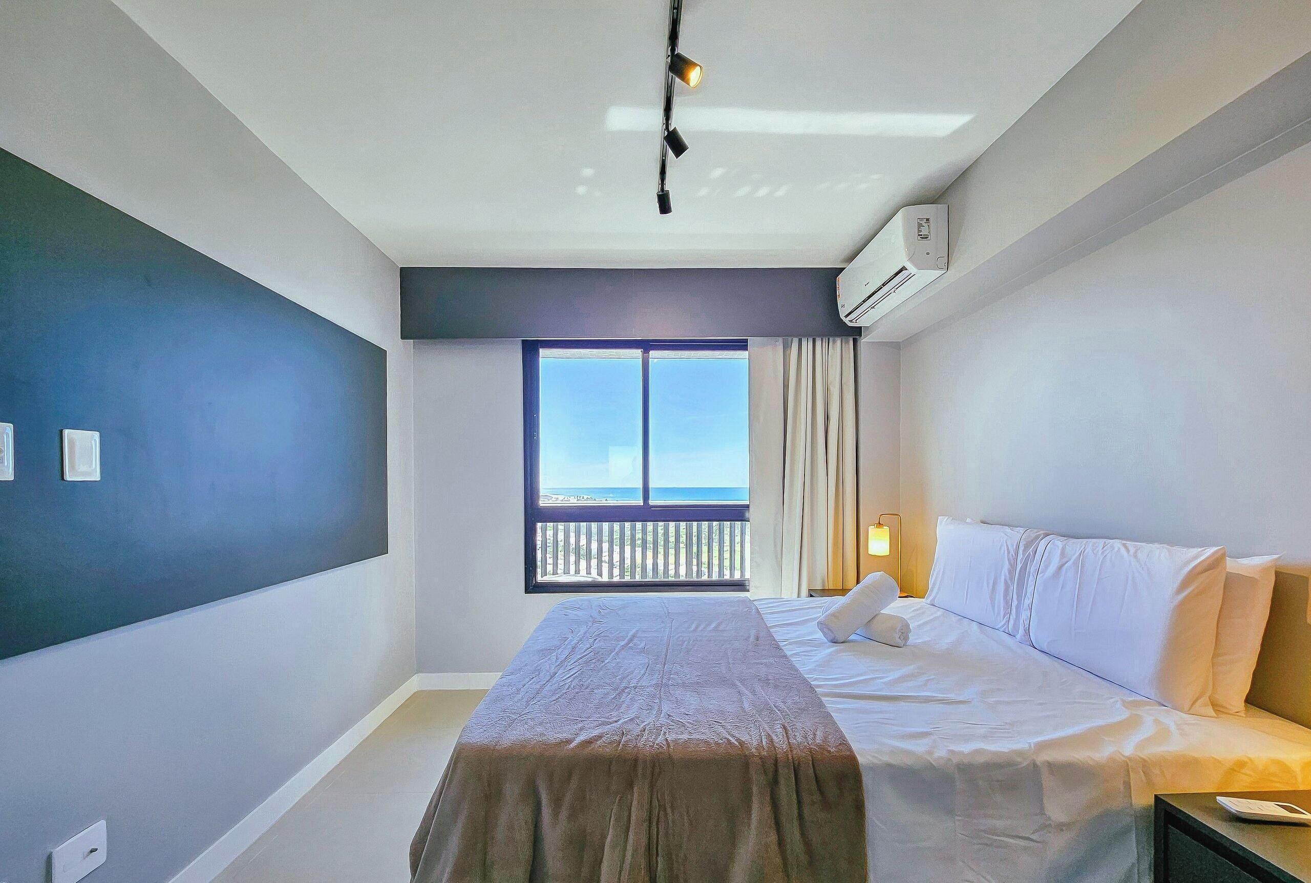 A hotel room with a bed, a window with a view, an air conditioning unit, and a ceiling light fixture.