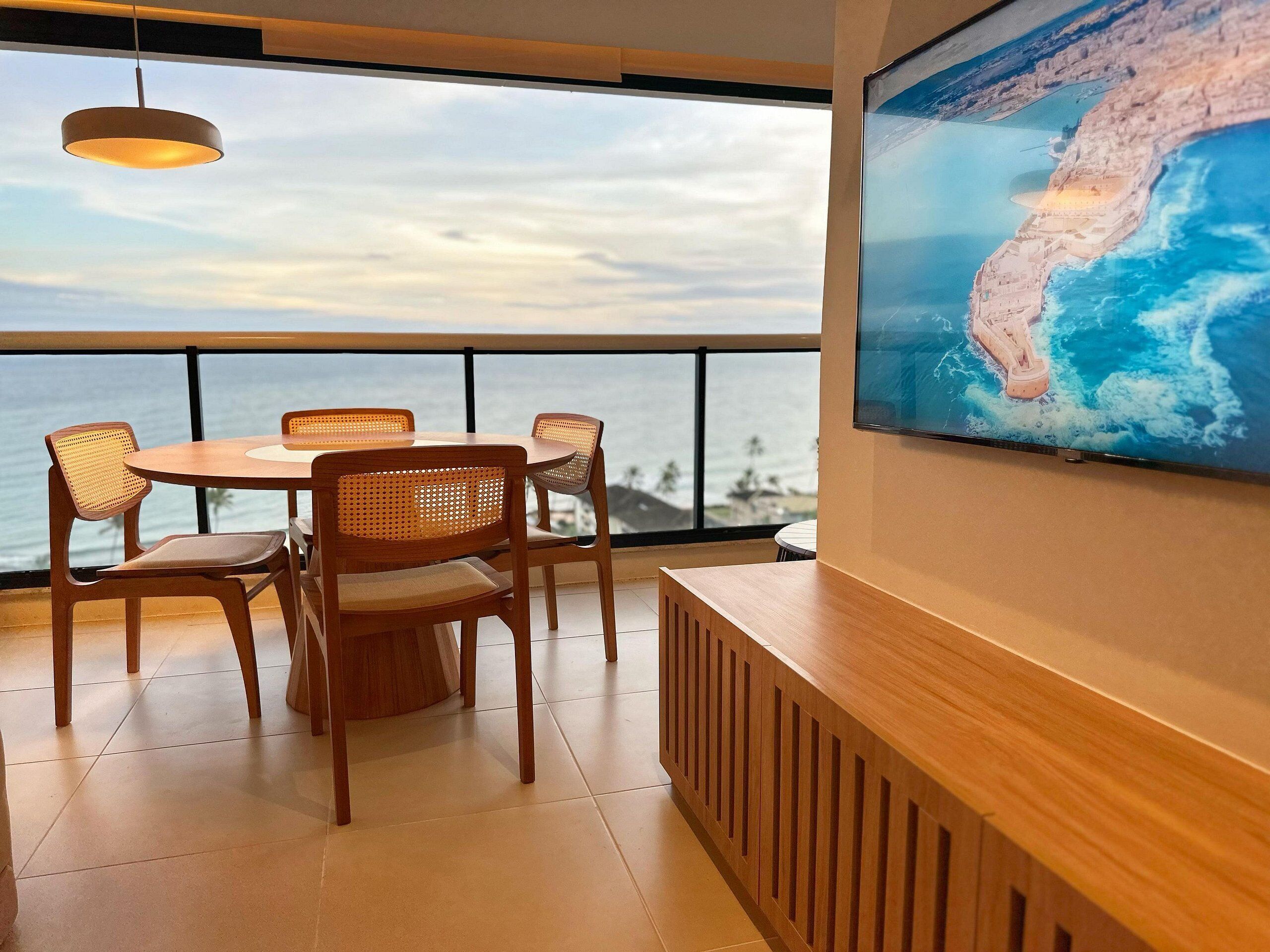 A dining area with a round table and four chairs, a hanging light fixture, and a large wall-mounted TV displaying a map.