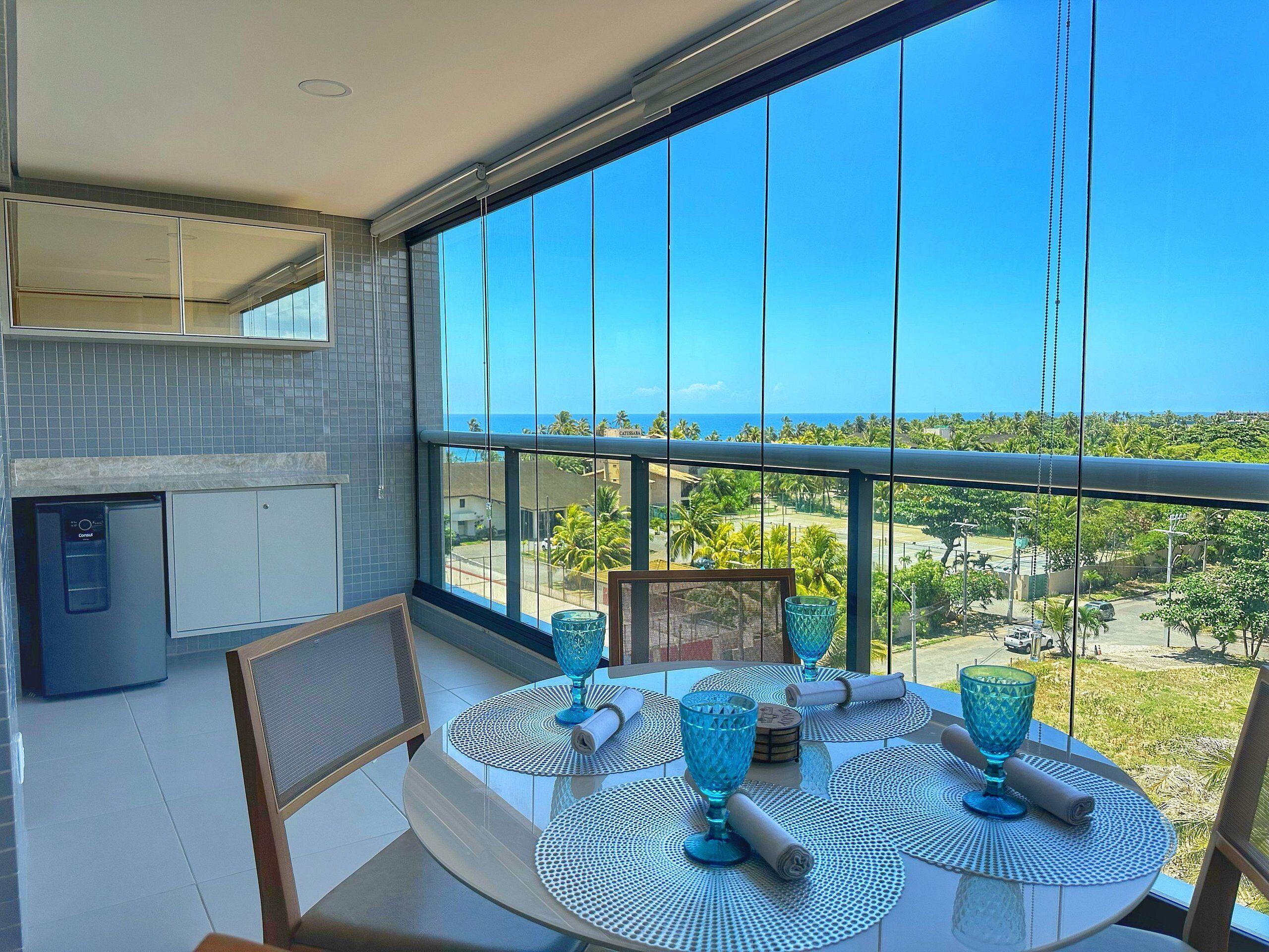 Un balcon avec une table dressée pour les repas, un réfrigérateur et une vue sur l’océan et la végétation.