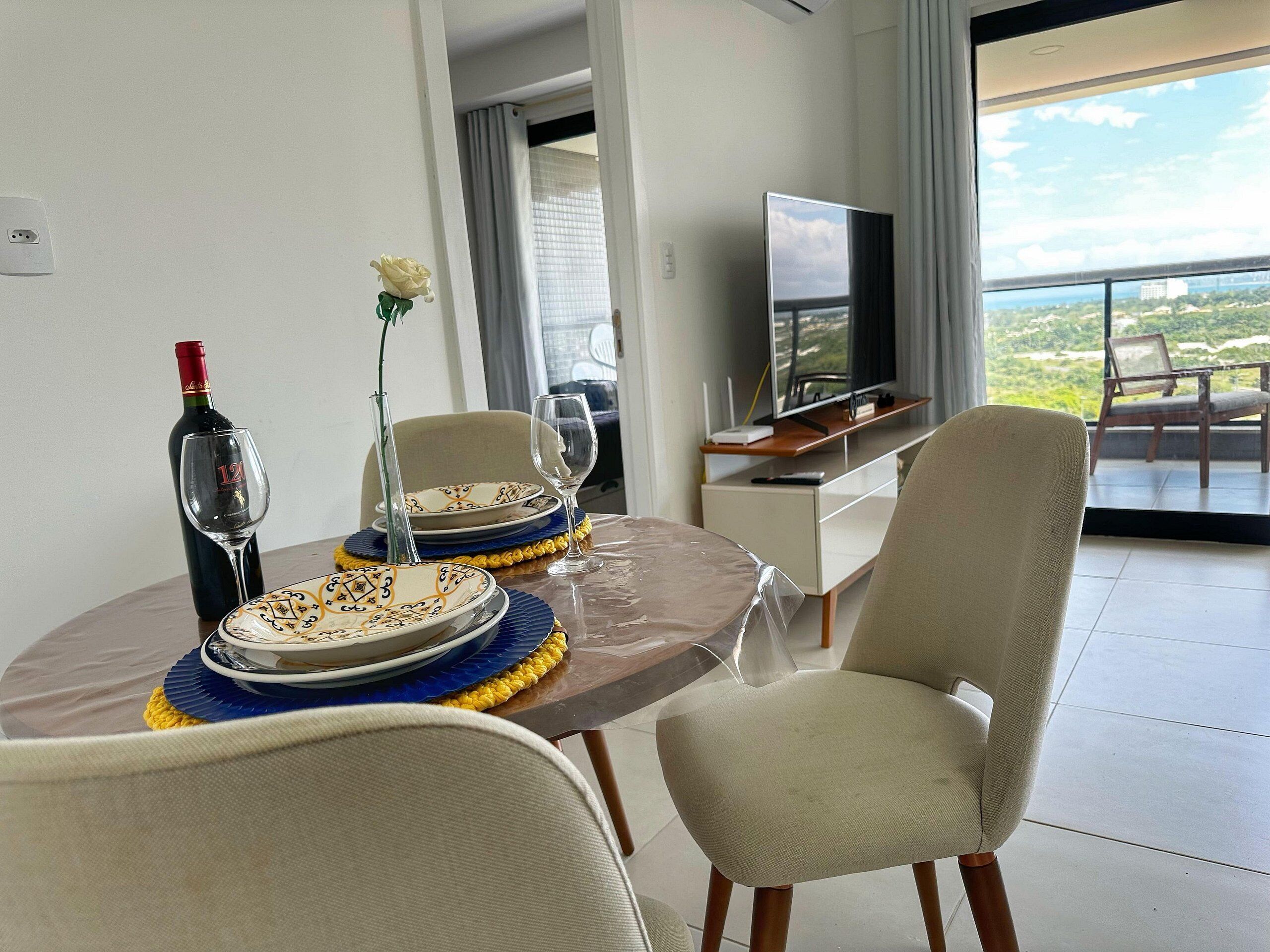 Una mesa de comedor con platos, una copa de vino y una botella, una silla, un televisor y un balcón con vistas.