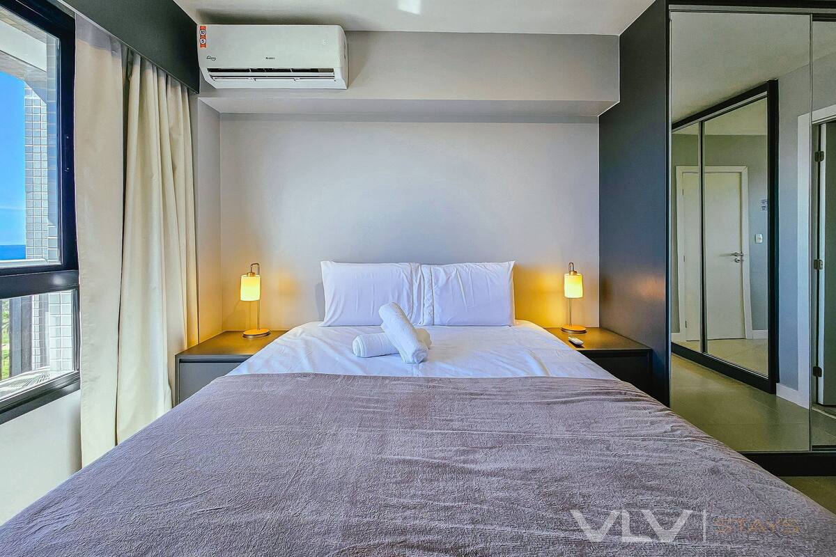 Una habitación de hotel con cama, dos lámparas de noche, un equipo de aire acondicionado, una ventana con cortinas y una puerta.