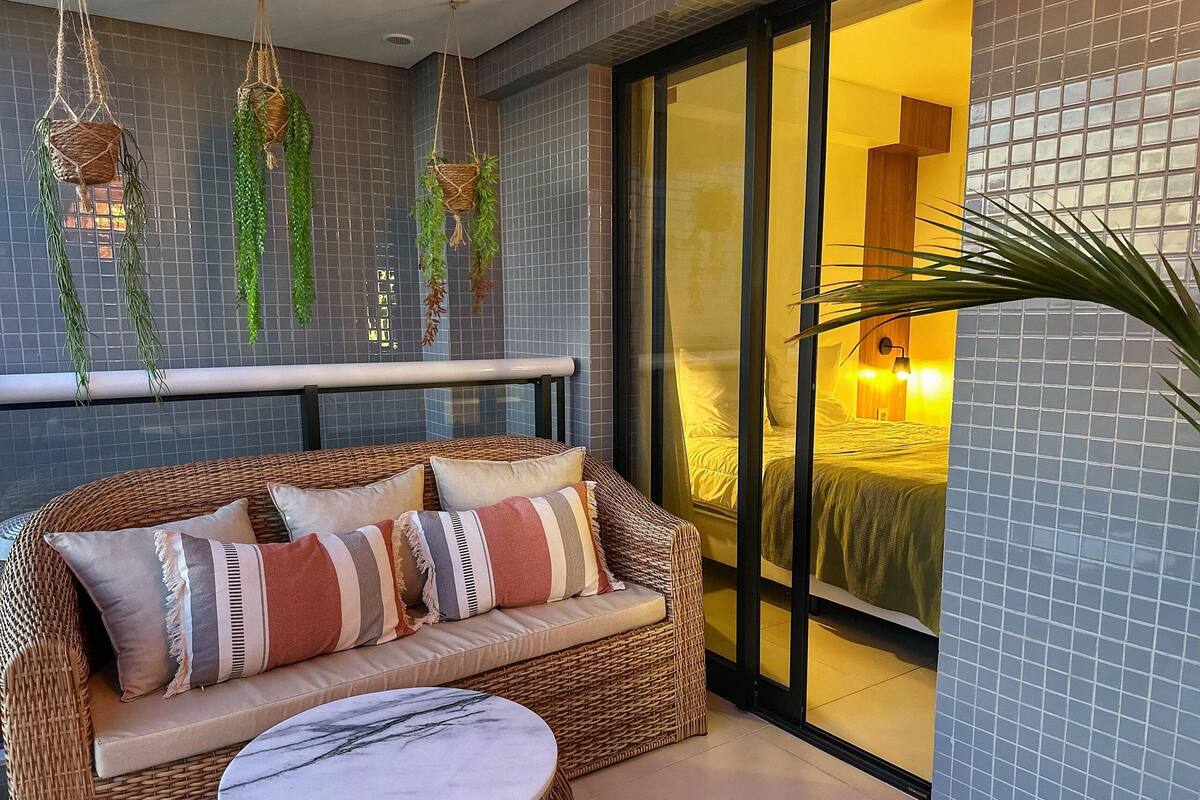 Un sofá de mimbre con cojines, una mesa de mármol, plantas colgantes y vistas a un dormitorio a través de una puerta de cristal.