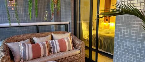 A wicker sofa with cushions, a marble table, hanging plants, and a view of a bedroom through a glass door.