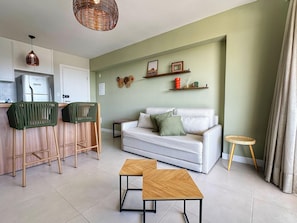 A modern living room with a white sofa, two green chairs, a small wooden table, and a kitchen area with a refrigerator. - Premium Stella Maris by VLV Stays (Salvador)