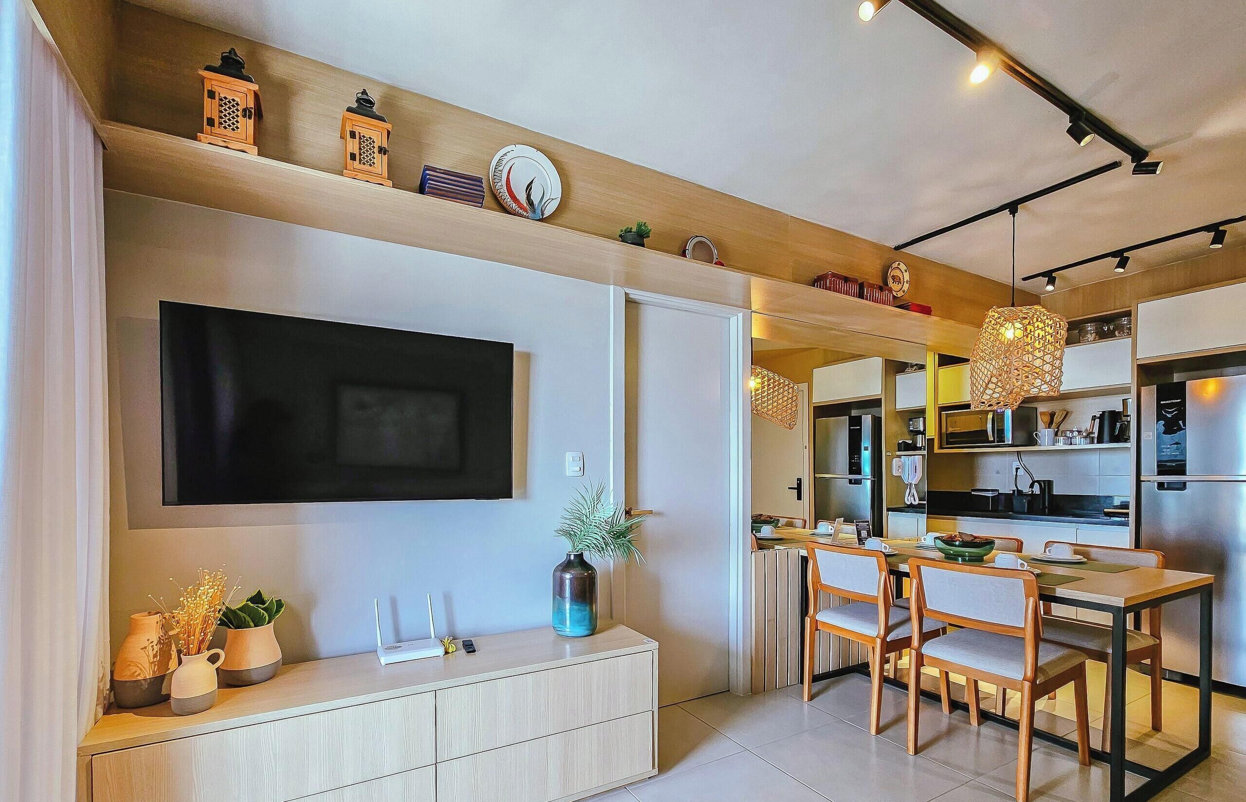 A modern living room with a flat-screen TV, a dining area with wooden furniture, and a kitchen with stainless steel appliances.