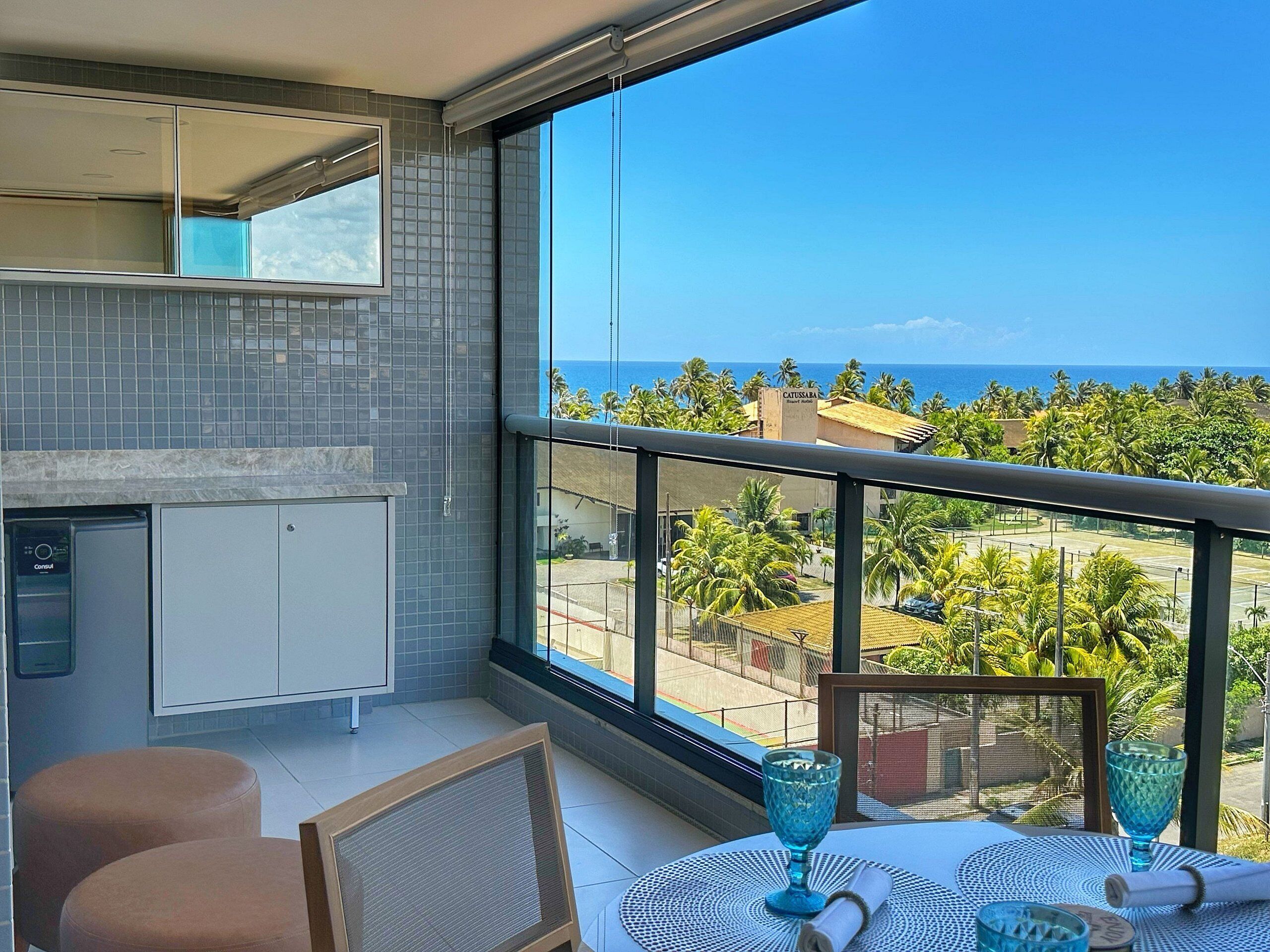 A balcony with a view of the ocean and palm trees, a dining table with blue glassware, and a kitchenette with a dishwasher.