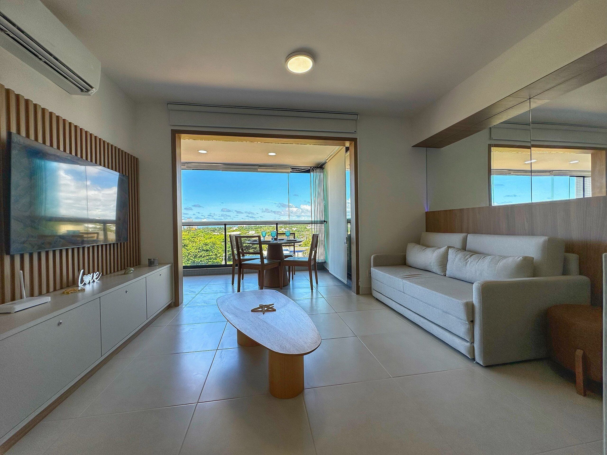 A modern living room with a flat-screen TV, a wooden coffee table, a sofa, and a balcony with a view.