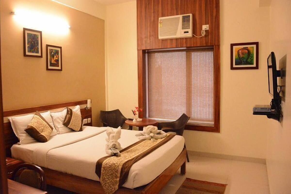 A hotel room with a large bed, a small table with chairs, a window with blinds, and a wall-mounted TV.