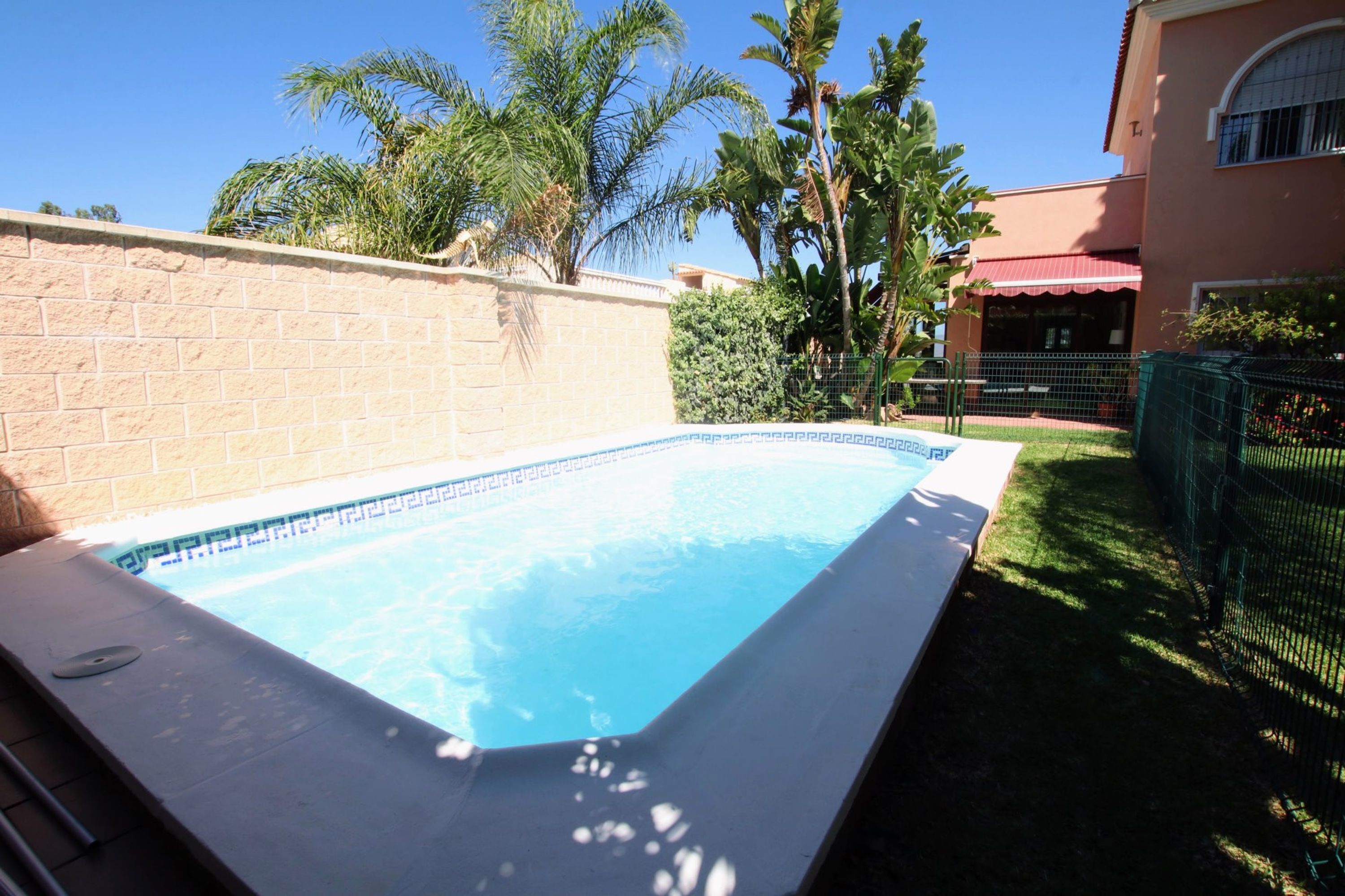 Une piscine rectangulaire dont le bord est recouvert de carreaux, entourée d’un mur de briques et d’une clôture verte.
