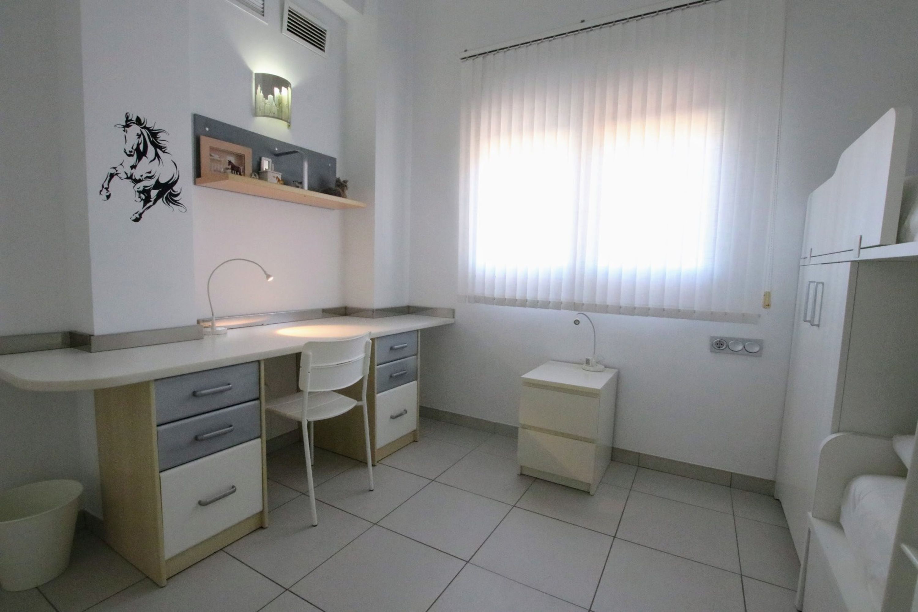Una habitación pequeña y limpia con escritorio, silla y ventana con persianas.