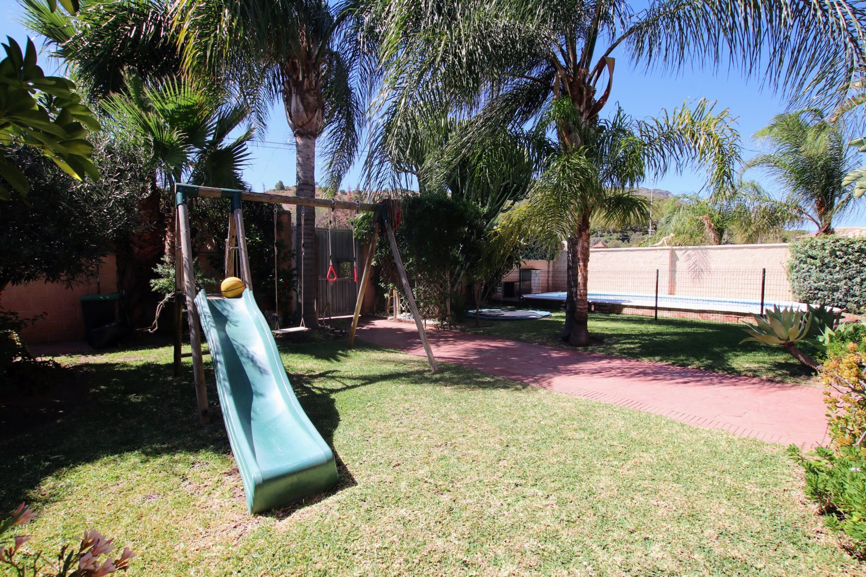 A playground with a slide and swings, surrounded by palm trees and a brick pathway.