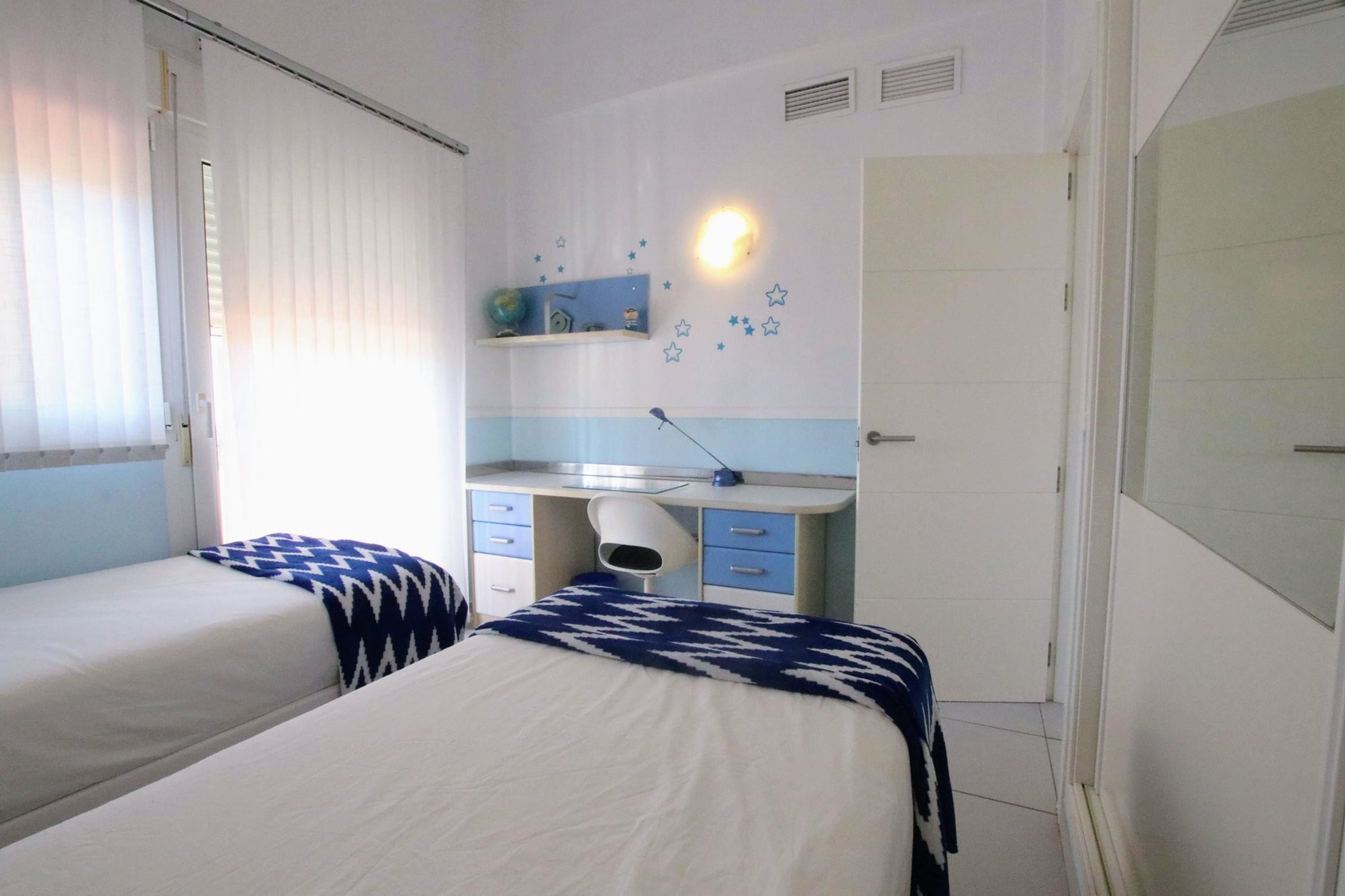 A bedroom with two beds, a desk, and a shelf with decorative items.