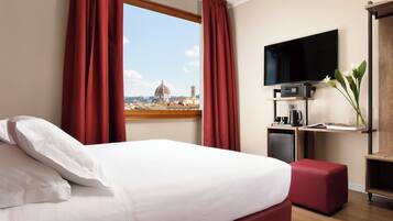 A hotel room with a bed, red curtains, a TV, a small desk with a coffee maker, and a view of a dome and cityscape.