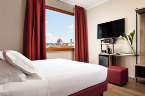 A hotel room with a bed, red curtains, a TV, a small desk with a coffee maker, and a view of a dome and cityscape. - Essence Hotel (Florence)