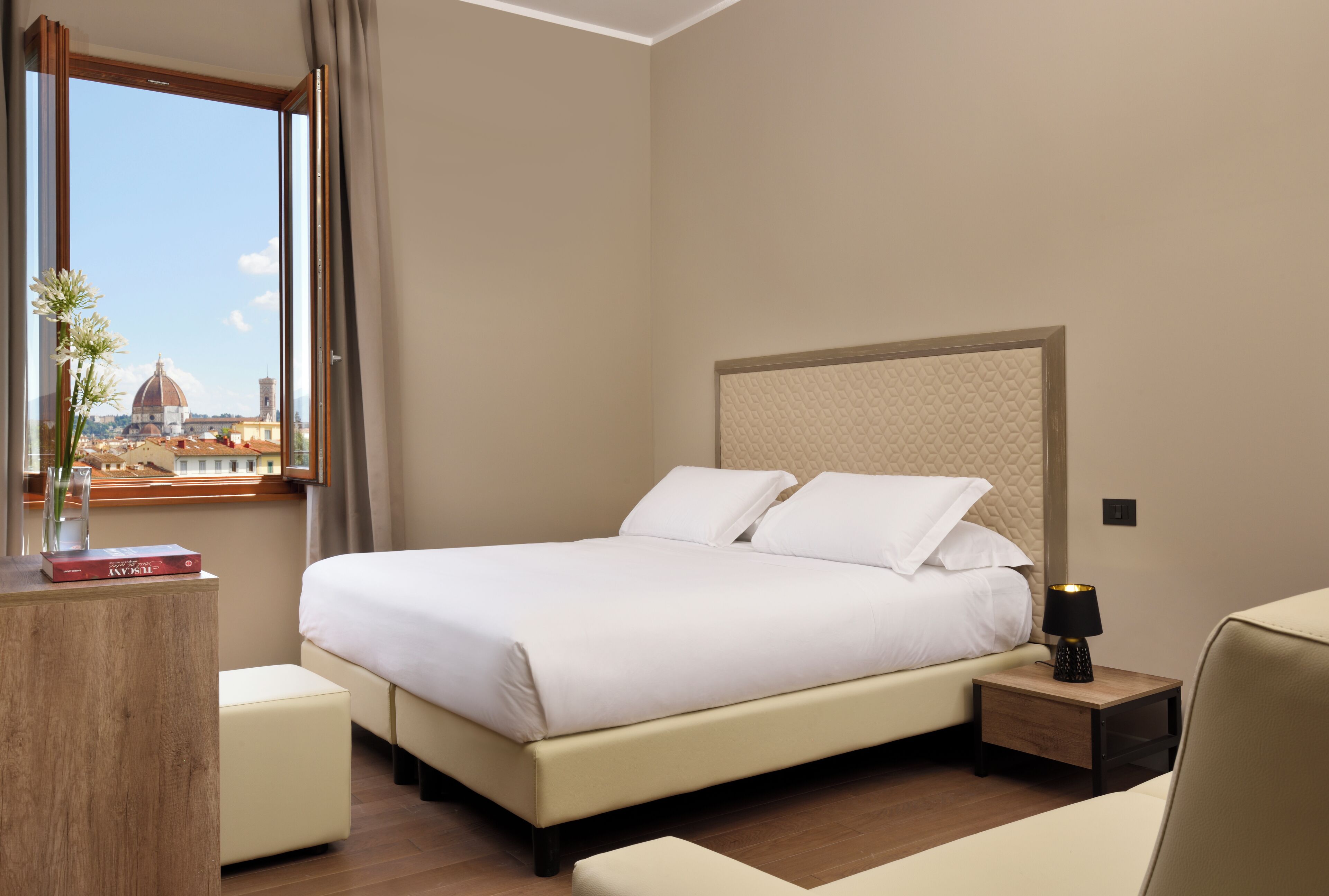 Hotelkamer met een bed, nachtkastje, lamp en raam met uitzicht op een koepel en gebouwen.