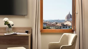 Hotelkamer met een groot raam met uitzicht op een koepel en gebouwen, een flatscreen televisie aan de muur, een houten bureau met een bloevas, een stoel en een klein poef.