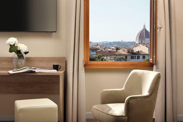 A hotel room with a large window offering a view of a dome and buildings, a flat-screen TV mounted on the wall, a wooden desk with a vase of flowers, a chair, and a small ottoman. - Essence Hotel (Florence)