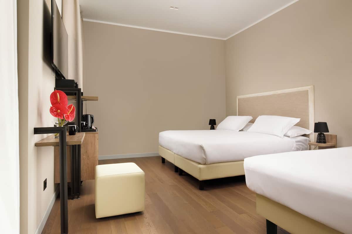 A hotel room with two beds, a wooden desk, a small stool, a TV mounted on the wall, and a nightstand with a lamp.