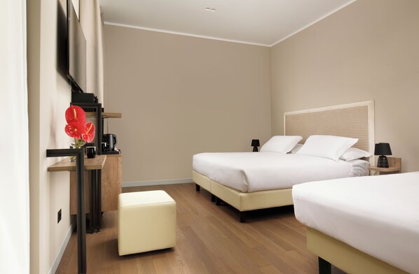 Ein Hotelzimmer mit zwei Betten, einem Holztisch, einem kleinen Hocker, einem an der Wand montierten Fernseher und einem Nachttisch mit Lampe.