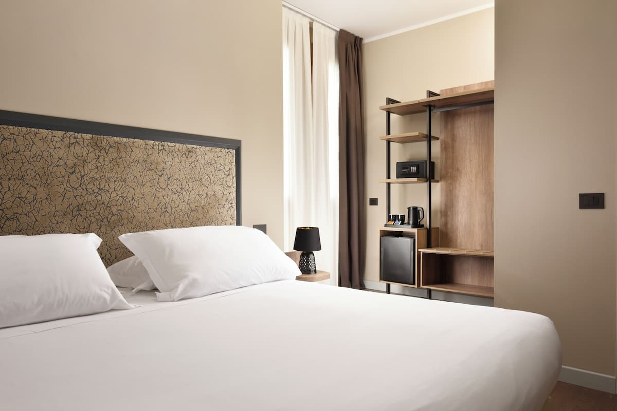 A hotel room with a large bed, a nightstand with a lamp, a wardrobe, and a small table with a coffee maker.