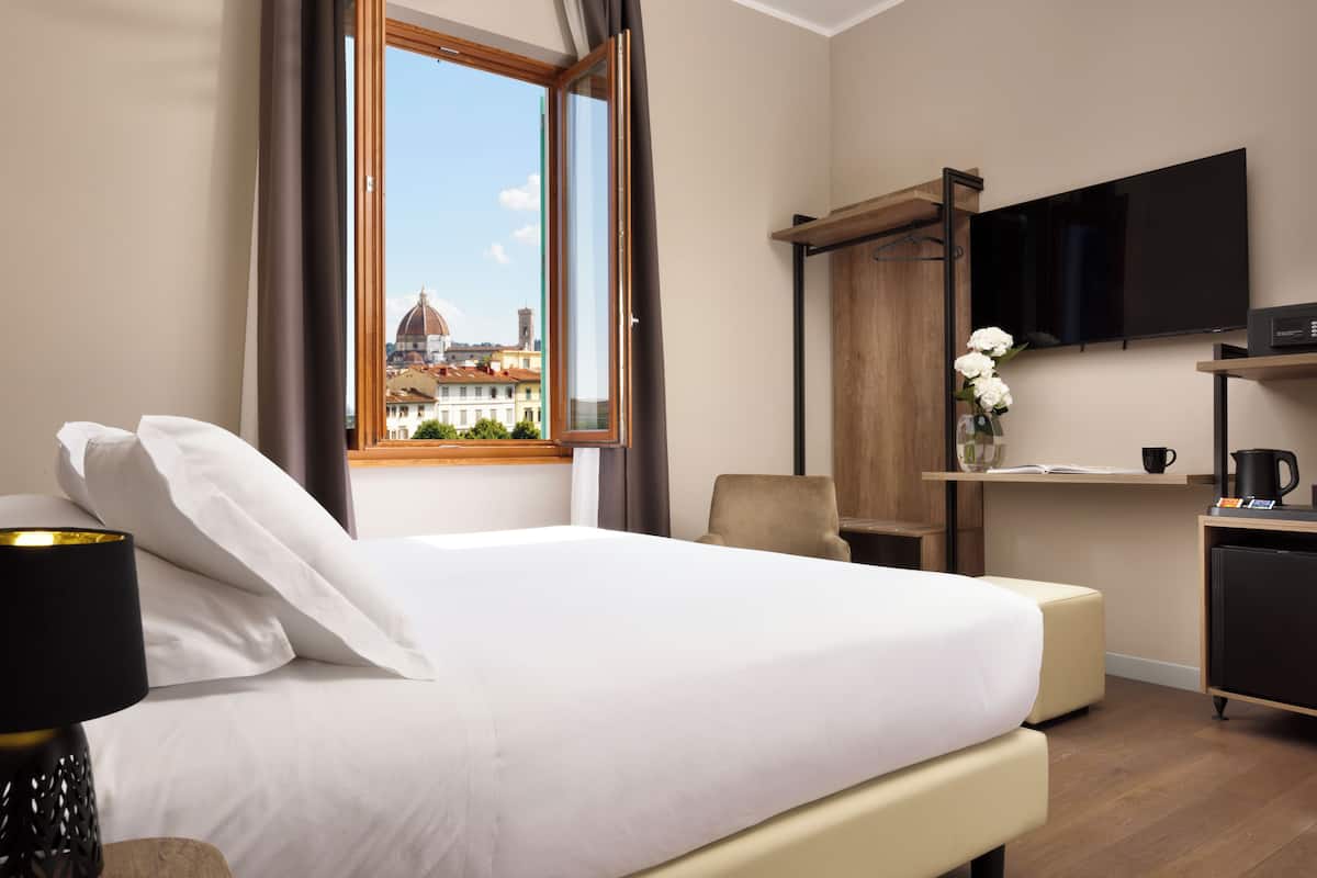 A hotel room with a bed, a desk with a flat-screen TV, a chair, and a view of a building with a dome.