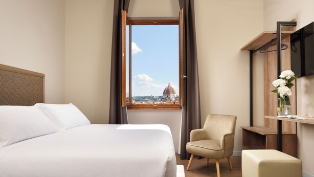Hotelkamer met een groot bed, een stoel, een tafeltje en uitzicht op een koepel door een raam.