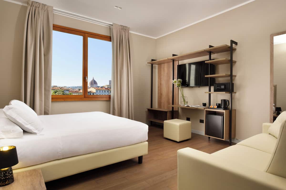 A hotel room with a bed, a desk with a TV, a coffee machine, and a view of a cityscape.