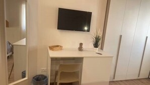 Un petit bureau blanc, avec un téléviseur fixé au-dessus, une chaise, une petite plante et une poubelle.