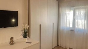 Une pièce dotée d’un téléviseur à écran plat fixé au mur, d’un meuble blanc orné d’une plante et d’une fenêtre agrémentée de rideaux légers.