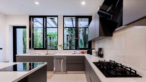 A modern kitchen with a central island, stainless steel appliances, and large windows offering views of greenery. - Villa Viola | Fully staffed New Luxury Villa | Laguna Golf Course & Beach Access (Pa Sak)
