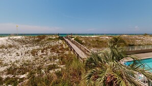 A beach view with a wooden boardwalk leading to the ocean, a pool area with lounge chairs, and a clear sky. - Gulf Dunes 102 3 Bedroom Condo by RedAwning (Fort Walton Beach)