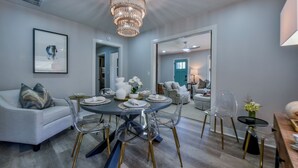 A dining area with a round table, clear chairs, and a chandelier. There is a sofa with pillows and a framed picture on the wall.