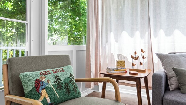 A cozy room with a large window, a chair with a hiking-themed pillow, a small table with books and a decorative item, and a grey sofa. - 5 mins to Acadia and 5 min walk to town! (Southwest Harbor)