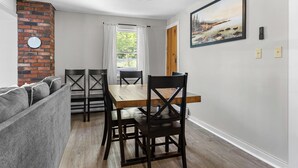 A dining area with a wooden table and chairs, a brick accent wall, a window with curtains, and a painting on the wall. - 5 mins to Acadia and 5 min walk to town! (Southwest Harbor)