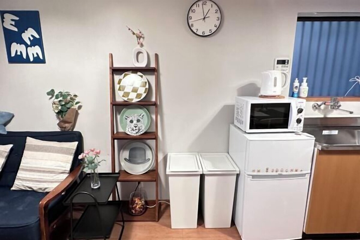 A compact living space with a sofa, a small table with a vase, a wall clock, a microwave, and a refrigerator.