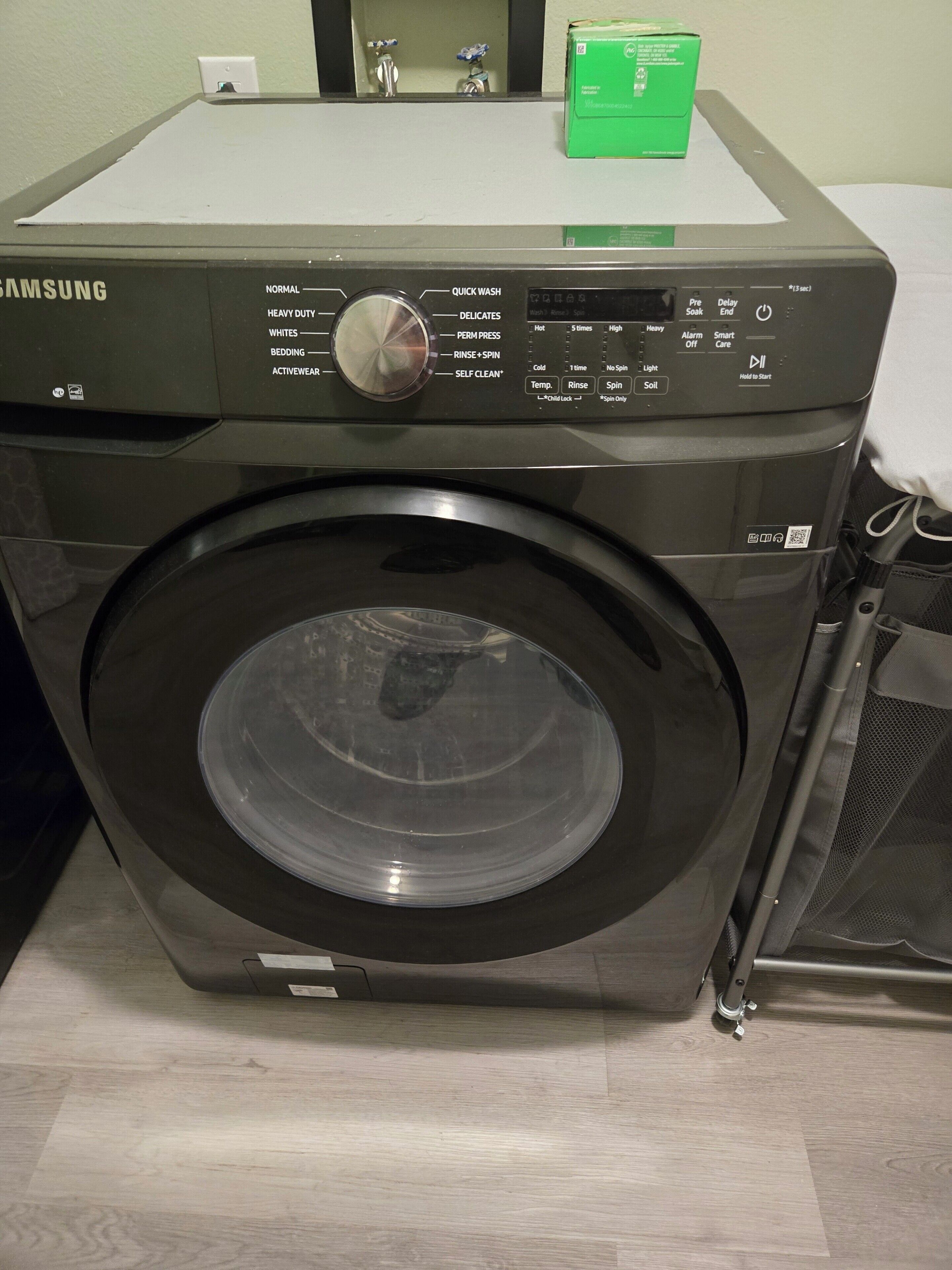 A Samsung front-loading washing machine with a control panel, a transparent door, and a green box on top.