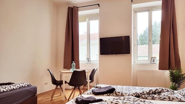 A hotel room with a bed, a desk, two chairs, a TV, and large windows with curtains. - Floridsdorfer Apartment (Vienna)